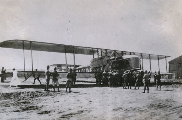 Three-quarter view from front right of a Lawson Air Liner parked near a hangar, with a group of men in military uniforms inspecting it. A woman in a white dress is walking next to the wing of the plane on the far left.