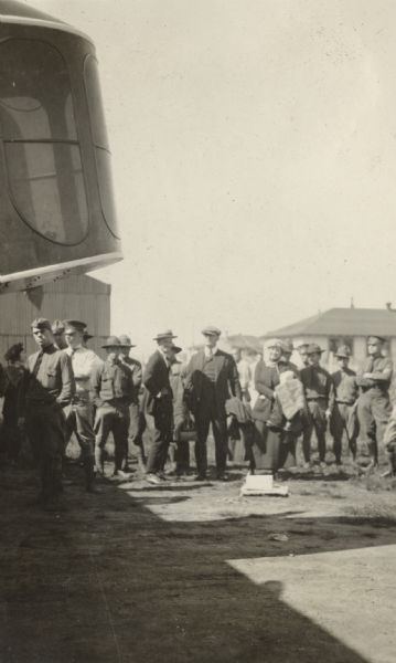 Several people are standing near a Lawson Air Liner. A number of people are wearing military uniforms, and others are wearing civilian clothes. Buildings are in the background. The cockpit of a Lawson Air Liner is  in front of the group.