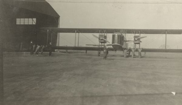 Men pushing on the wings of a Lawson Air Liner outdoors near a hangar.