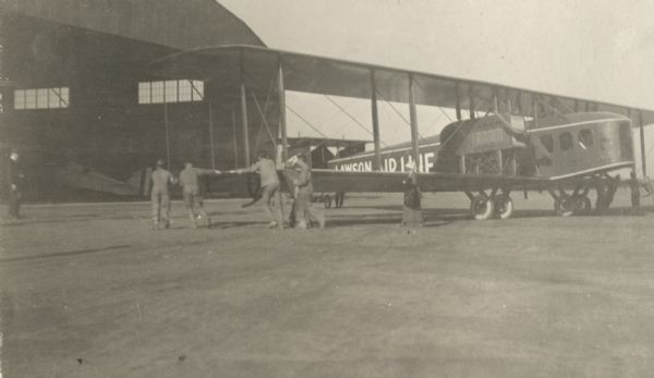 Several men are pushing a Lawson Air Liner by its wings back into an open hangar. A woman is walking near the front of the wing.