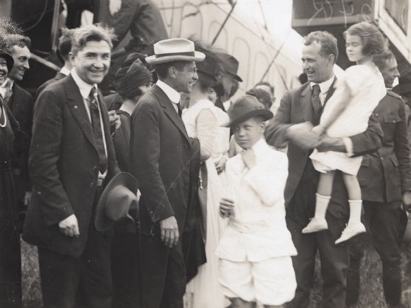 Alfred Lawson standing and talking to United States Secretary of War Newton Baker. Lawson is holding Baker's daughter Betty in his arms, and Baker's son is standing between them. A crowd of people are in the behind them, and a Lawson Air Liner is in the background. This image was taken on the event of a flight Lawson gave around Washington D.C.