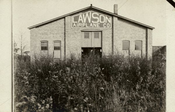 Exterior view from field with tall plants towards one of the entrances to a factory building with the Lawson Airplane Company sign over it.