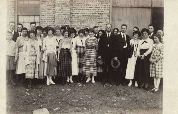 Outdoor group portrait of Alfred Lawson posing with large group of women, and a few men. They are standing in front of what appears to be the Lawson Airplane Company factory.