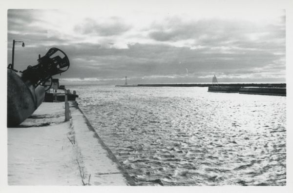 View from jetty out towards Lake Michigan entrance to Ship Canal. A large tank, streetlamp and buildings are on the left. On the opposite jetty is a lighthouse. Snow is on the ground.