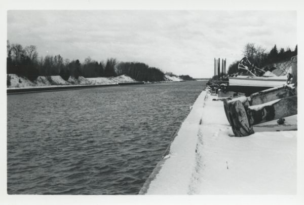 View of the Ship Canal from the Coast Guard station. There are pieces of machinery on the right, and a line of trees on the left. There is snow on the ground.