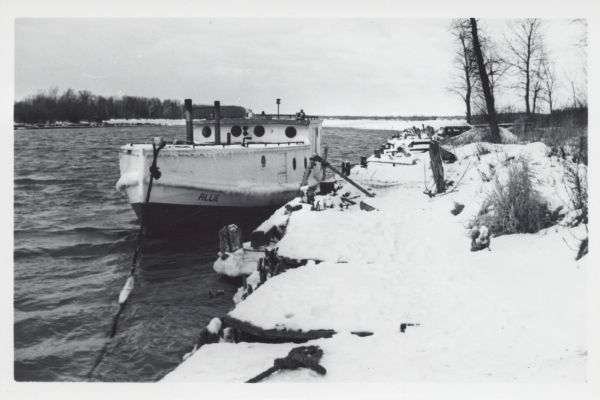 View from snowy shoreline of Mooring Basin at bay end of Ship Canal. The boat moored in the foreground is named "Allie." It is tethered to a post on the shoreline and is layered with icicles. Across the bay is a forested shoreline.