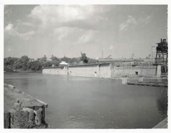 View across water towards the Manitowoc Shipbuilding floating drydock. Pilings and grass are in the foreground. On the far shoreline are buildings on a hill among the trees. Machinery and industrial buildings are on the right, including multiple cranes. The floating drydock is made of concrete.