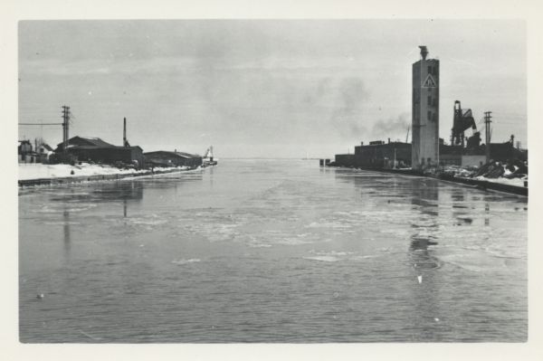 Elevated view from 8th Street Bridge over the Manitowoc River towards Lake Michigan. There is a lighthouse in the distance. Ice is floating on the water. Along the left and right shorelines are industrial buildings and materials.