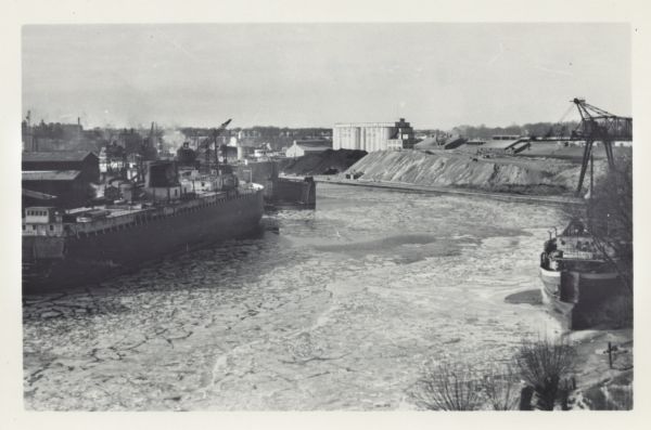 Elevated view over icy water towards ships in the shipyard on the left, and the cement company in the background. Cranes and industrial buildings are in the background. Another ship, with name on front reading "Marquis Roen," is moored along the shoreline in the right foreground.