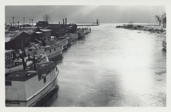 View from the Second Street bridge over the icy Ahnapee River towards Lake Michigan. A lighthouse is at the end of a jetty in the far background. Boats are moored along the left shoreline.