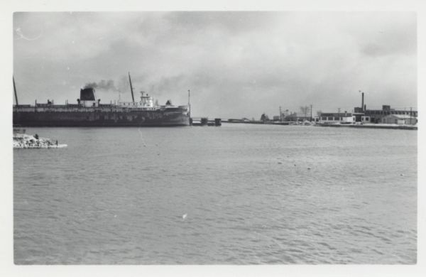 View across water to ship moored at a dock. Smoke is coming out of the smokestack. Industrial buildings are across the water on the right.