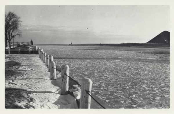 View of Two Rivers Harbor from shoreline towards Lake Michigan. Snow is on the ground, and a fence made of thick posts and cable is along the shoreline on the left, with a lighthouse in the far background. The river is frozen. On the opposite shoreline is a large pile of what may be coal.
