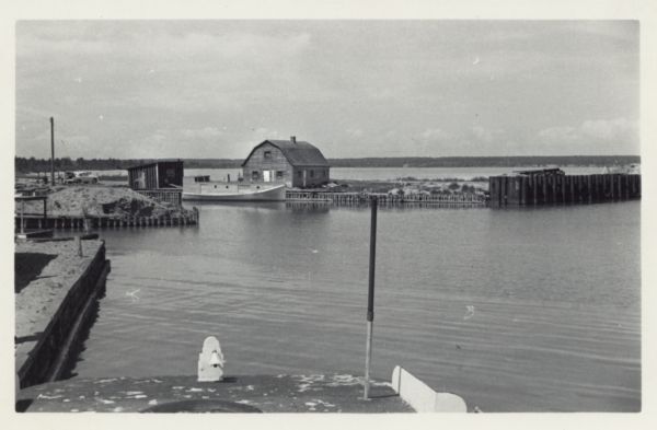 Slightly elevated view of Cornucopia Harbor. There is a boat moored at a pier near a barn-shaped building. An automobile is parked on the far left. In the background is the forested shoreline across the bay. In the foreground is a bell on top of what may be a boat roof.