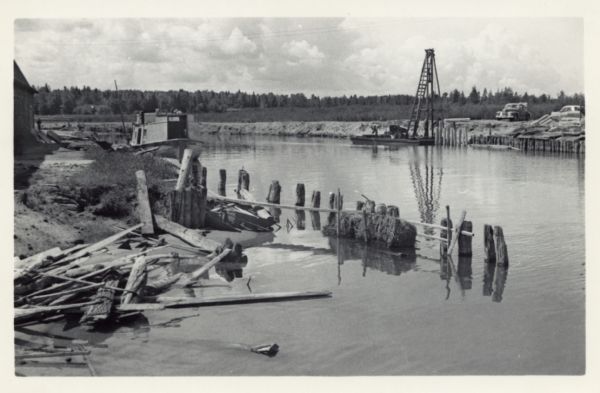 View over water towards storm damage in Cornucopia Harbor. West slip. There is a pile of wood along the shoreline in the foreground, with a boat moored just behind near a building on the far left. On the right shoreline men are on a raft with a tall wooden apparatus, perhaps for installing piling. Two automobiles are parked nearby.