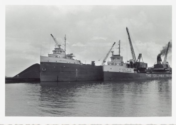 View across water towards ships unloading coal and sulphur. Cranes rise above both ships, and a man is walking near a large pile of coal in the background on the left. The ship on the left is named: "David J. Norton," and the ship on the right is named: "W.C. Richardson." Along the water line of the ship are the words: "The Columbia Trans... ."