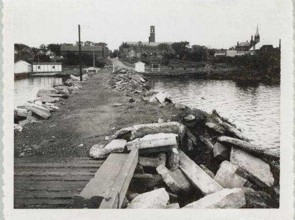 View from pier, lined with rocks, towards town. There are industrial buildings along the shoreline, and up a small rise is a building with a tower, and on the far right is a church building with a steeple.