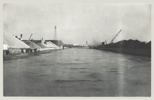 View across water, with cranes among piles of coal along the right, and among piles of other materials on the left. Smoke is in the distance on the right, and large power lines and storage tanks are on the left.