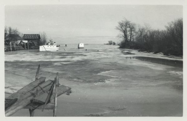 View across icy water towards shipping boats in harbor. Trees are along the right shoreline, houses and buildings are along the left shoreline.