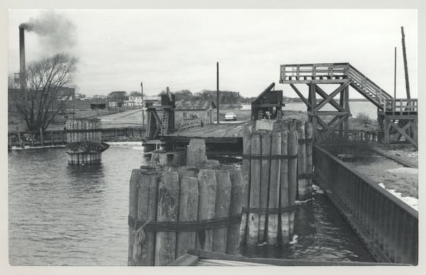 Carferry slip with pilings in the foreground, and a shoreline in the background. The pier is made of timbers. In the background on the left is a factory with a tall smokestack.