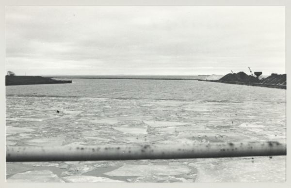 View of icy Lake Michigan from Ogden Street Bridge. There are piles of coal on the left and right, and what may be a jetty in the far distance.