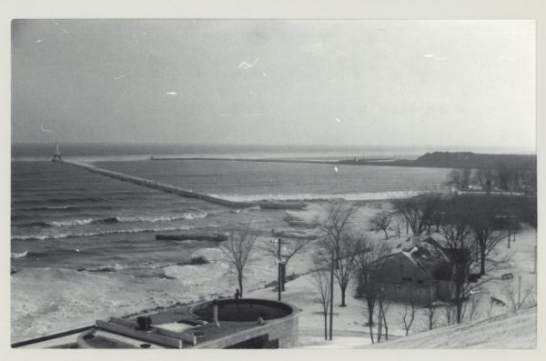 Elevated view from bluff of the harbor towards the southeast. At the end of a jetty is a lighthouse on the left. The water is frozen along the shoreline. There are industrial buildings in the foreground.