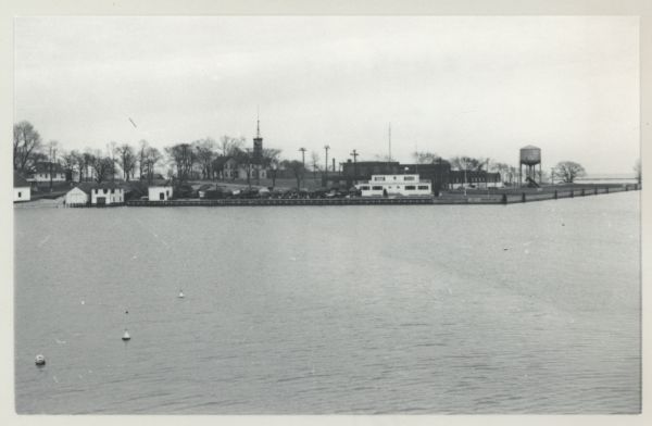 Turning basin, view east from ex-Haas property. There is a water tower on the right, and houses, boathouses and other buildings are along the shoreline to the left. Covered boats are stored above the water on the wharf.