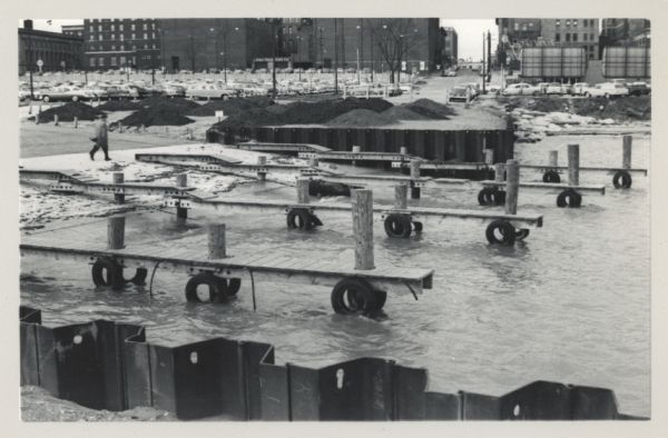 Pleasure boat ramps, made of timber, on the south side of Racine Harbor. A man is walking in the snow near the ramps. There are piles of material along the wharf near the empty boat ramps. In the background is a parking lot near large brick buildings.