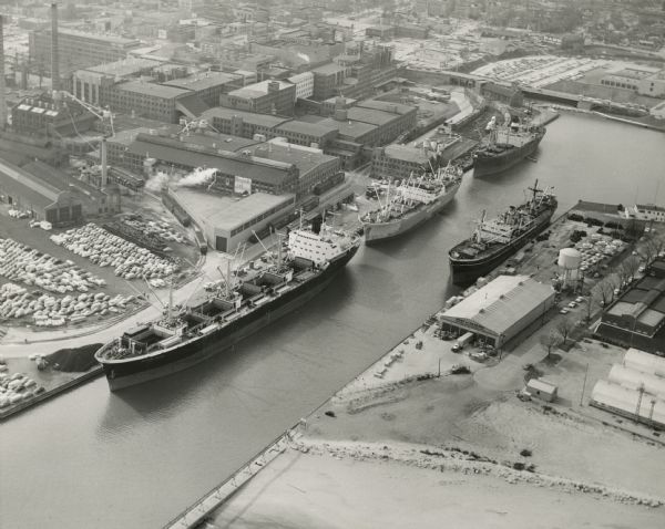 Aerial photograph of the Port of Kenosha and factories, with snow on the ground. There are four ships along the shoreline, and multiple factories on either side. A building on the right has a sign that reads: "Port of Kenosha." On the left is a parking lot with cars.