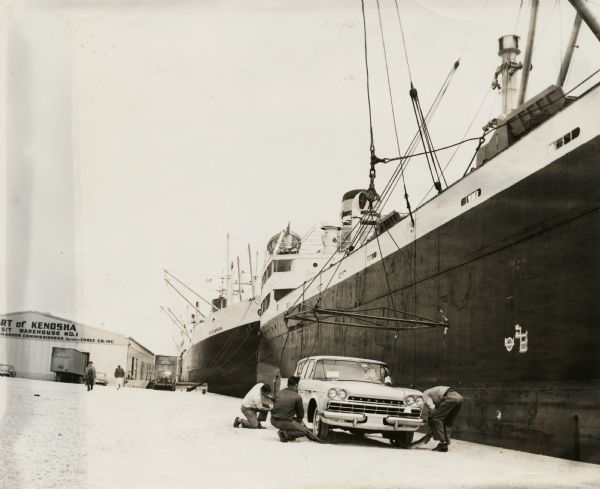 Port of Kenosha Transit Warehouse No. 1. There are two ships on the right side, one named "Broom Park." Three men are loading a car onto the ship using a crane. Snow is on the ground.