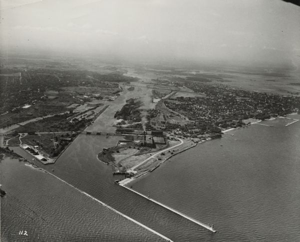 Aerial view of harbor. In the foreground is a lighthouse and breakwaters. Bridges cross the harbor, and there are factory and industrial buildings near the shorelines. On the right and left are trees, streets, and houses.