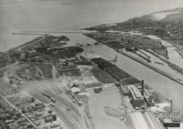 Aerial view of Marinette Harbor looking out towards Lake Superior. A lighthouse and breakwater extend out into the lake. Industrial buildings, cranes, and railroad tracks are along the shoreline.