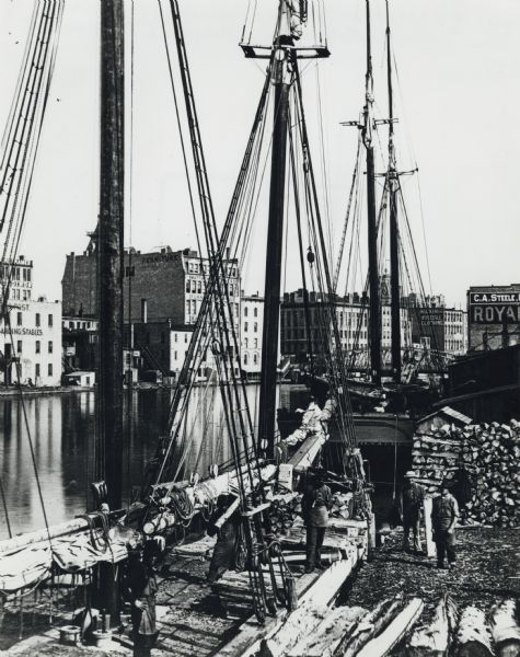 Elevated view of a deck of a schooner hauling lumber. Several workmen are loading or unloading the lumber. Industrial buildings, some displaying signs, are on the opposite shoreline. In the background is a bridge.