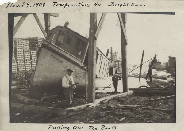 Men working on pulling the boat "Fish Hawk" out of the water. One man is kneeling in front of the boat and looking at the camera. A man in the background is using a pole to move the boat. On the far right a boy is standing in a boat watching. A stack of pallets are stacked in the left background. Handwritten on top: "Temperature 40. Bright Sun," and on the bottom: "Pulling Out the Boats."