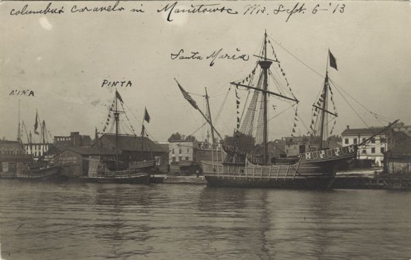 Postcard view across water towards reproductions of Christopher Columbus' ships, the <i>Niña</i>, <i>Pinta</i>, and the <i>Santa Maria</i> docked at Manitowoc. The ships, adorned with flags and banners, are probably docked along the Manitowoc River.