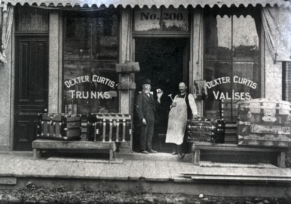 View from street of two men standing just inside the open doorway of the storefront. Trunks are displayed on pallets set up on the wooden sidewalk. Dexter Curtis, the inventor of the horse collar pad and a manufacturer, operated this business at 206 E. Main Street. In a 1901 directory, his business is listed as "trunks and collar pads."