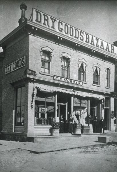 View from street of the C.E. Morgan Dry Goods Bazaar on North Pinckney at East Mifflin. Two women are standing in the doorway on the left, and two men are standing in the doorway on the right. In 1908, the goods in the store were assigned to Edwin Coy 'for the benefit of the creditors.'