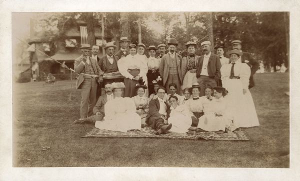 Group portrait of the Edward M. Fuller family and guests during a pause at a 'frolic' at the residence located east of the Yahara River. Part of the group is standing in front of and behind a hammock attached to two tree trunks. Another group is sitting in front on a carpet spread on the lawn. There is a house in the background with a large U.S. flag draped from an upper window.