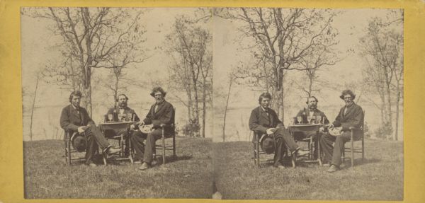 Three men posing while sitting in chairs at a table with trees and a lake in the background. On the table are glasses and a bottle on a tray.