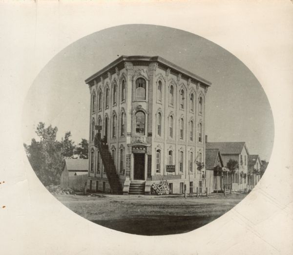 Oval-framed view from street of the corner bank building at 111 S. Hamilton Street, with houses on the left and right. The main business is Millers Bank Insurance. It is also occupied by the Jas. Richardson and Co. Land Office and several law offices, including C. Abbott Law Office. It is the largest and most imposing building on the block. There is a long iron stairwell that leads from the sidewalk up to a second story entrance on the left side of the building, where two men are posing. The trees on the street appear to be newly planted and have fences around them. Manoah D. Miller, a Baptist minister, built this bank about 1857. He closed the bank in 1861. The building is no longer at 111 S. Hamilton and Main Street, near S. Carroll Street. It was replaced by the Jackman Building which was built in 1913.