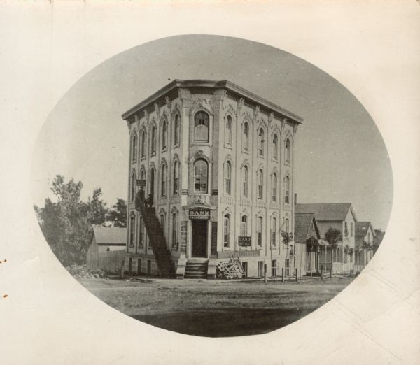 Oval-framed view from street of the corner bank building at 111 S. Hamilton Street, with houses on the left and right. The main business is Millers Bank Insurance. It is also occupied by the Jas. Richardson and Co. Land Office and several law offices, including C. Abbott Law Office. It is the largest and most imposing building on the block. There is a long iron stairwell that leads from the sidewalk up to a second story entrance on the left side of the building, where two men are posing. The trees on the street appear to be newly planted and have fences around them.

Manoah D. Miller, a Baptist minister, built this bank about 1857. He closed the bank in 1861. The building is no longer at 111 S. Hamilton and Main Street, near S. Carroll Street. It was replaced by the Jackman Building which was built in 1913.