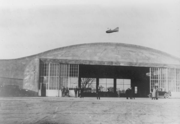 The Royal Airways Corporation Hanger at the airport on Monona Drive, built in 1929. Among those standing in front are E.N. Quin, President; Howard Morey, Vice-President; G.W. Stephans, Secretary; Al Breuch, Treasurer. On the right side, in front of the glass hangar doors is a small runabout automobile. On the left a child is standing next to one of the men. There is a small blimp attached to a pole on the roof of the building.