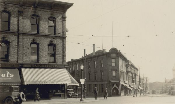 View from street towards the H.H. Ratcliff Company, jewelers and opticians, at 29 S. Pinckney Street. Across the street, the building on the corner of Main Street (to the left) and King Street (to the right) was built for John J. Suhr and designed by John Nader. The first floor of this building is used by "mechanical stores,' which has mounted a large calipers above the entrance door. On the left side of the mechanical stores is a shoe store. There is a streetcar coming up King Street.
