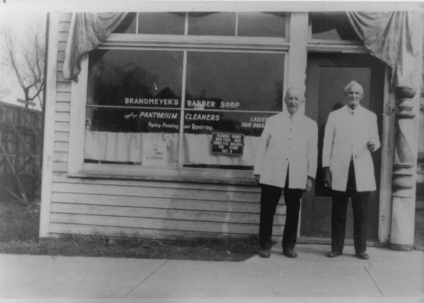 Two barbers standing and posing on the sidewalk in front of the James J. Brandmeyer Barber Shop at 404 W. Lakeside Street. A sign in the window advertises that they are the agent for Pantorium Cleaners, dyeing, pressing and repairing. They also do Ladies Hair Bobs. Another sign states their closing hours and another one that says the Alderman is T.P. Connor.