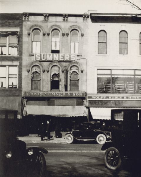 View across street towards the Edwin Sumner and Son Drugstore at 15 S. Pinckney Street. Automobiles are parked at an angle on both sides of the street, and people are standing on the sidewalk in front of the drugstore. There was an earlier store on Carroll Street and a later store, owned by his son L.D. Sumner, on State Street. Edwin Sumner opened his drugstore in 1859, and until a celebration of sixty years in business in 1919, never closed a single day. Next to Sumner's is the Wolff, Kubly, Hirsig shop, which also had multiple locations over time on the square.