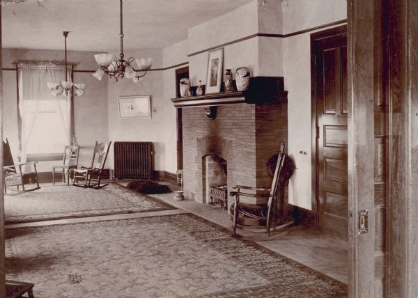 Interior view of a living area through a pocket door in the Chi Psi fraternity at 627 North Lake Street and Lake Lawn Court. The house was built in 1892 and occupied by the fraternity until 1909. There are worn rugs on the floor and several rocking chairs. The fireplace has been used for a fire. There are knob light switches on the wall beside a door on the right. On the floor near the fireplace, is a spitoon, and also a sheepskin or bearskin rug.