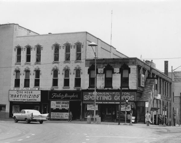 View from street of businesses along West Main Street, long after the remodeling of the Blied Building, 29 West Main Street, when the top two stories were removed. Businesses shown are Forbes-Meagher Music Store at 27 W. Main Street, and Wisconsin-Felton Sporting Goods. There is an automobile passing in front of the One Hour Martinizing shop and pedestrians are walking along Hamilton Street. The Forbes Meagher store has a sign in the window saying they have moved to 112 N. Fairchild. The Wisconsin-Felton Sporting Goods store is having a moving sale.