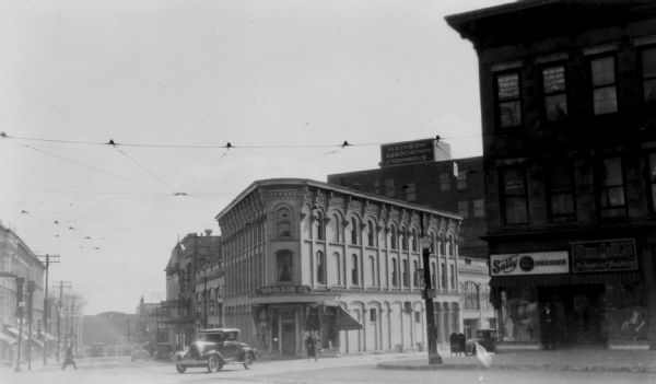 View from street towards the corner of King Street and S. Pinckney Street. On the corner is the Haw-Olson Company. Down King Street is the Majestic Theater and the Frautschi Furniture Company. There is a streetcar coming up the hill on King Street. Across Pinckney Street on Main Street are Sally Dresses and Rundell's Men's Clothing Store.