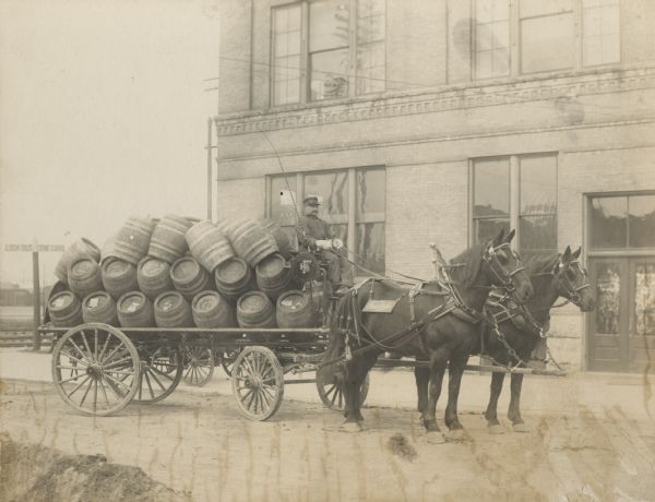 Joseph Bossart driving a horse-drawn wagon loaded with 37 empty beer barrels is parked in front of the International Harvester building on S. Blount Street near Williamson Street, built in 1898. The ends of the barrels have stamps that read: "Fauerbach BRG CO, Madison, WIS." A sign hanging from the harness on the horse reads "Fauerbach." In the background on the left are railroad tracks and a sign that reads: "Look Out for the Cars."
