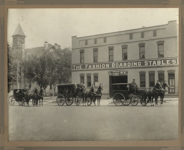 View across street towards the Fashion Boarding Stables, also called The Fashion Livery, located at 219-223 E. Washington Avenue on the corner at E. Butler Street, which was owned by E.S. Burwell. There are three men, each on a horse-drawn carriage, posing in the street in front of the stables. In the background on the left is a church building.