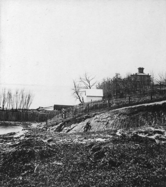 View along shoreline with Lake Monona on the left. A fence runs along the slope, and beyond is a small white building and behind, at the top of the hill, Farwell's octagonal house. The house was completed by Governor Farwell in 1854. In January of 1866 it was converted to the Soldiers' Orphans Home. The house stood on an entire block bounded by Brearly, Lake Monona and Spaight Streets. The home was razed in 1895. [Hanks describes this as 'Pump Carpenter's.']